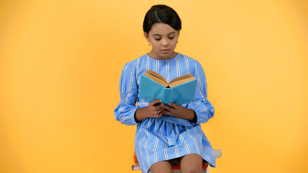 školačka v modrých šatech čtení knihy, zatímco sedí izolované na žluté - Fotografie, Obrázek
