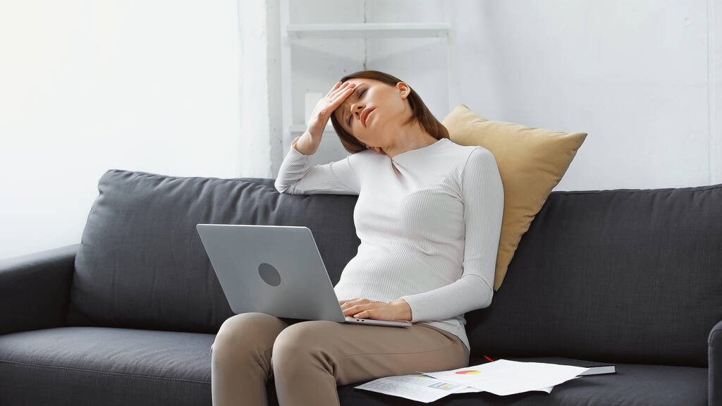 agotada, mujer embarazada que sufre de dolor de cabeza cerca de la computadora portátil y papeles en el sofá - Foto, imagen