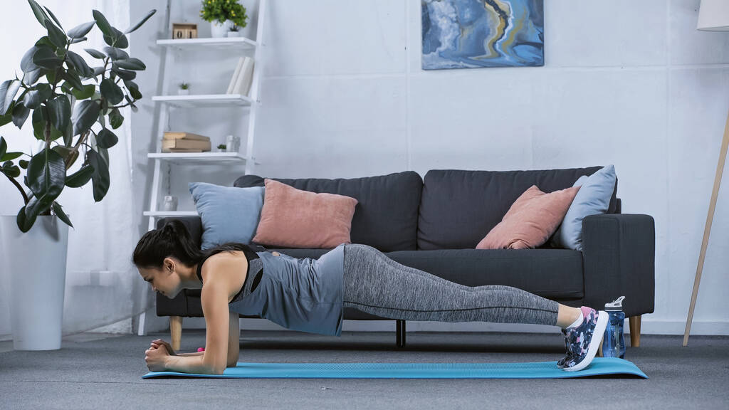vue latérale de la jeune femme en tenue de sport faisant planche sur tapis de fitness à la maison  - Photo, image