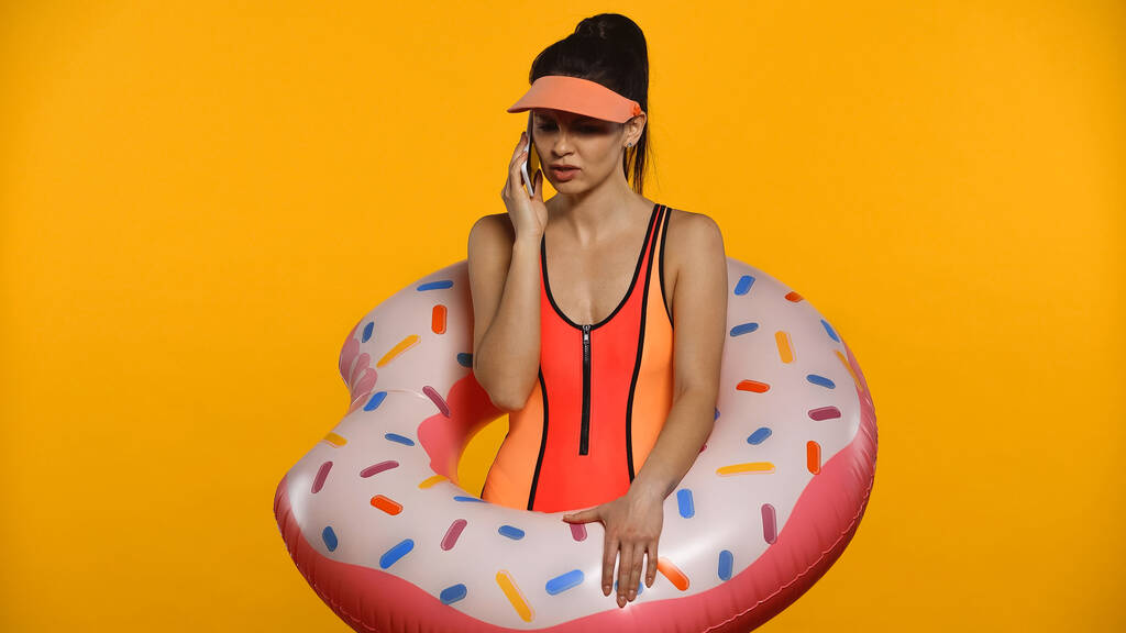 upset young woman in swimsuit and inflatable ring talking on smartphone isolated on yellow - Photo, Image