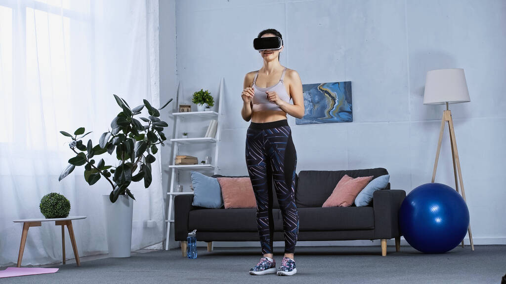 jovem mulher em sportswear e vr fone de ouvido em pé na sala de estar moderna - Foto, Imagem