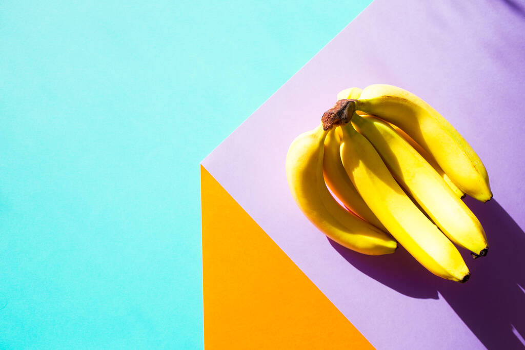 φύλλα χαρτιού δημιουργούν ψευδαίσθηση τρισδιάστατου κύβου. ένα μάτσο μπανάνες με σκληρή σκιά. γεωμετρικό φόντο σε φωτεινά χρώματα - Φωτογραφία, εικόνα