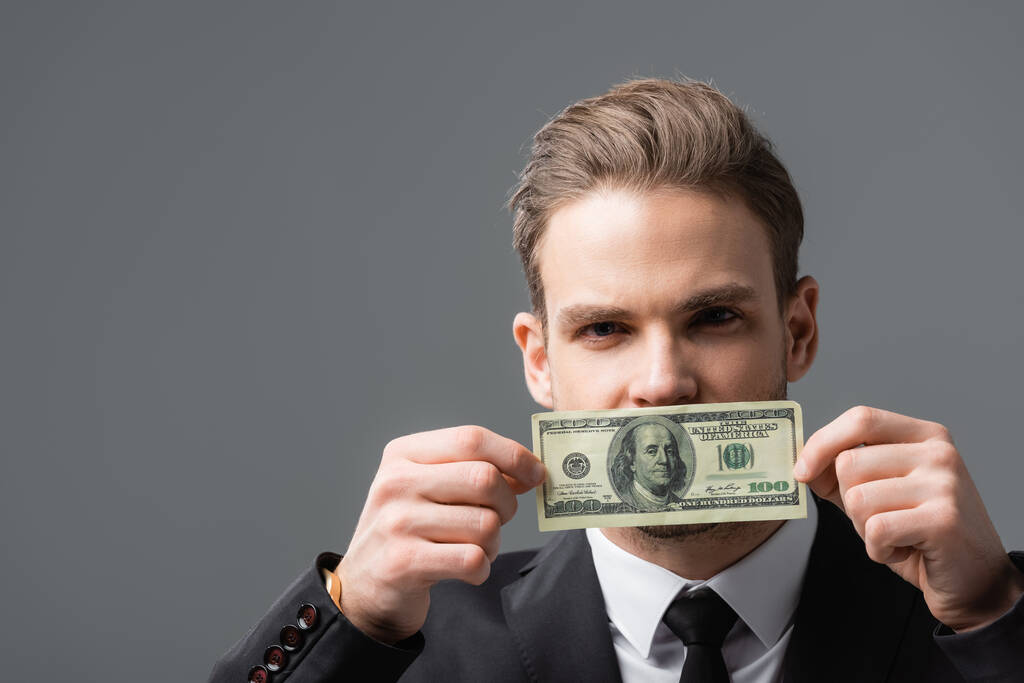 giovane uomo d'affari che copre la bocca con banconote in dollari isolate su grigio - Foto, immagini