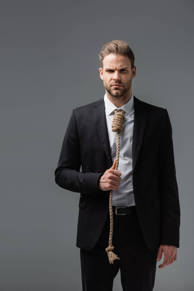 gespannen zakenman in zwart pak, met touw in plaats van stropdas, kijkend naar camera geïsoleerd op grijs - Foto, afbeelding