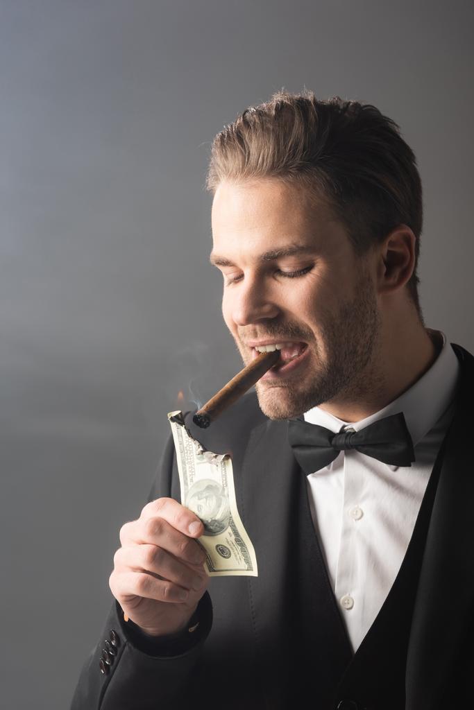 богатый бизнесмен зажигает сигару со стодолларовой банкнотой на сером фоне с дымом - Фото, изображение