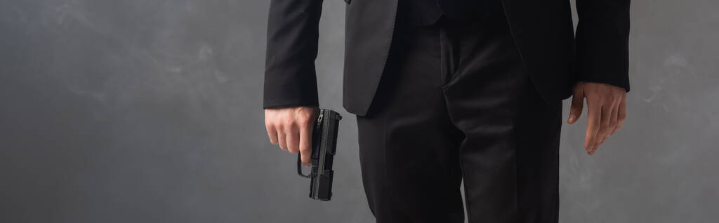 partial view of businessman in black suit holding weapon on grey background with smoke, banner - Photo, Image
