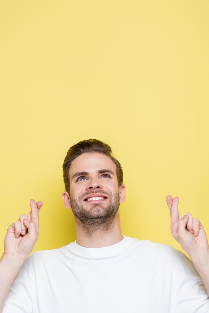 homem positivo segurando dedos cruzados enquanto olha para cima no amarelo - Foto, Imagem