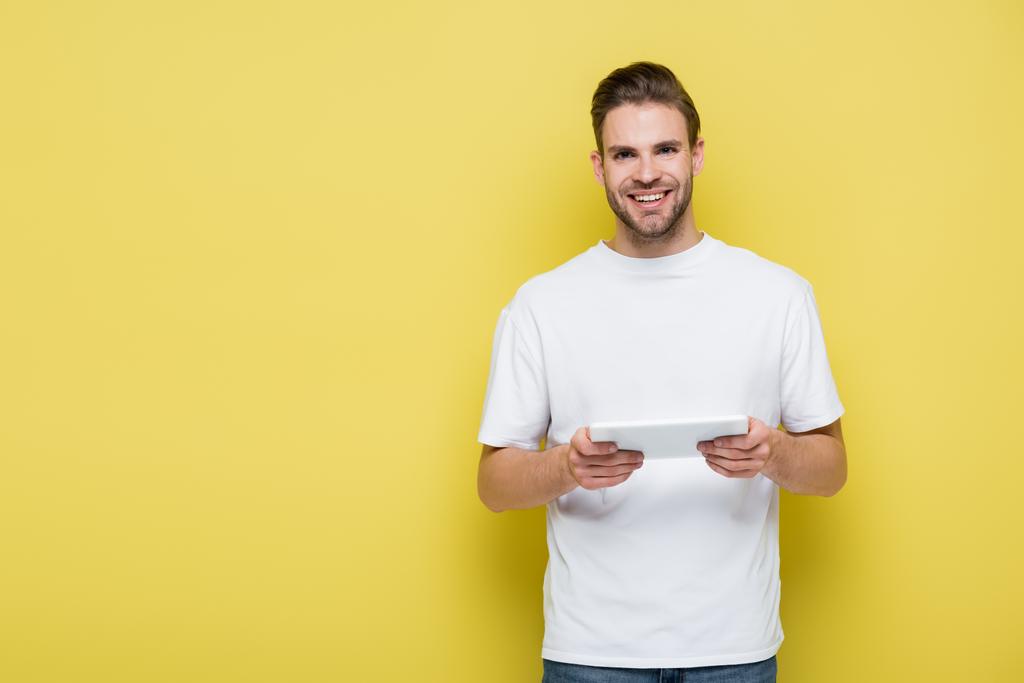 boldog férfi fehér pólóban kezében digitális tabletta és mosolygós kamera sárga - Fotó, kép