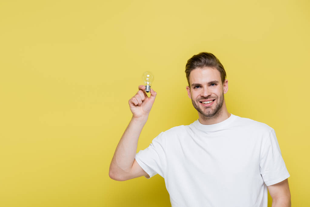 alegre hombre sosteniendo la bombilla mientras sonríe a la cámara en amarillo - Foto, Imagen