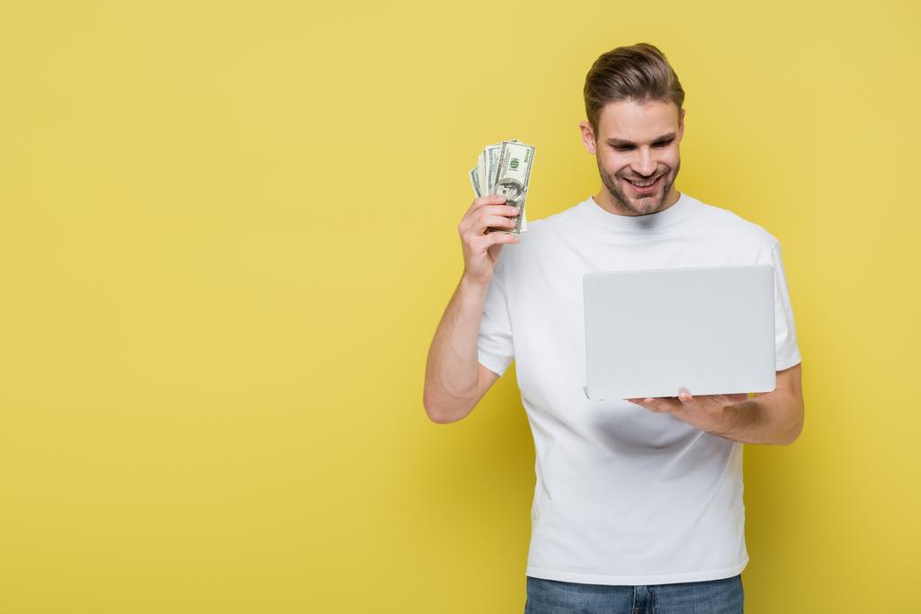 довольный мужчина держит долларовые банкноты, глядя на ноутбук на желтом - Фото, изображение