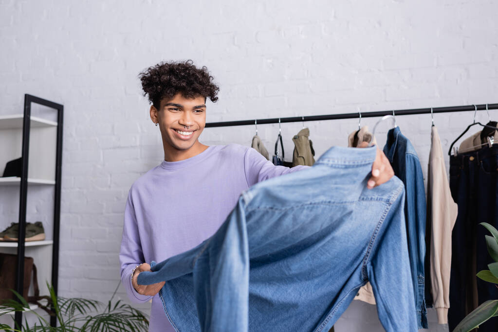Fröhlich afrikanisch-amerikanischer Showroom-Besitzer hält Kleiderbügel mit Jeanshemd auf verschwommenem Vordergrund  - Foto, Bild