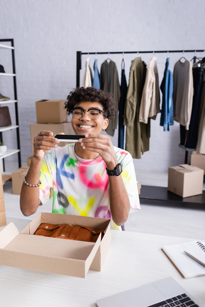 Glimlachend Afrikaans-Amerikaanse eigenaar van showroom het nemen van foto 's op smartphone in de buurt van doos met kleren en notebook  - Foto, afbeelding