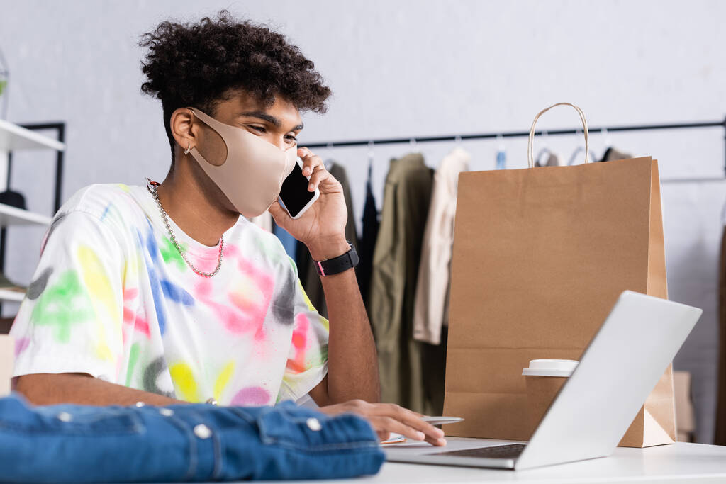 Africano americano proprietário de showroom em máscara protetora falando no smartphone e usando laptop perto de saco de compras  - Foto, Imagem