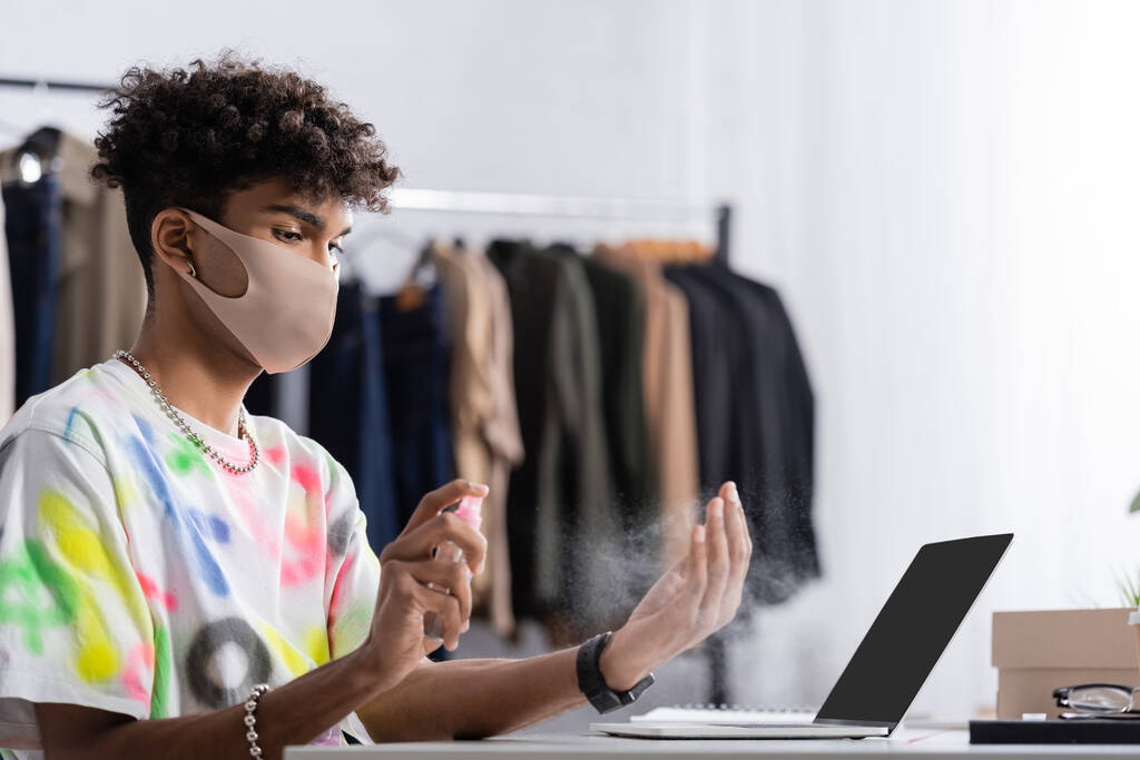 African american small business owner in protective mask spraying hand sanitizer near laptop  - Photo, Image