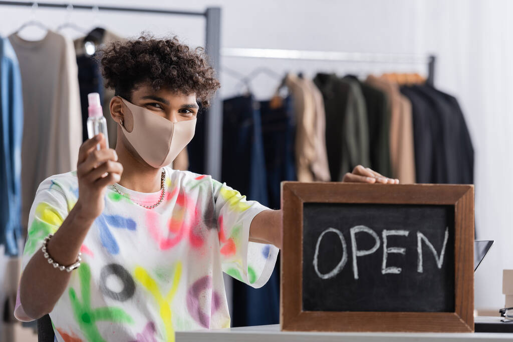 African american owner of showroom in protective mask holding hand sanitizer and chalkboard with open lettering on blurred foreground  - Photo, Image