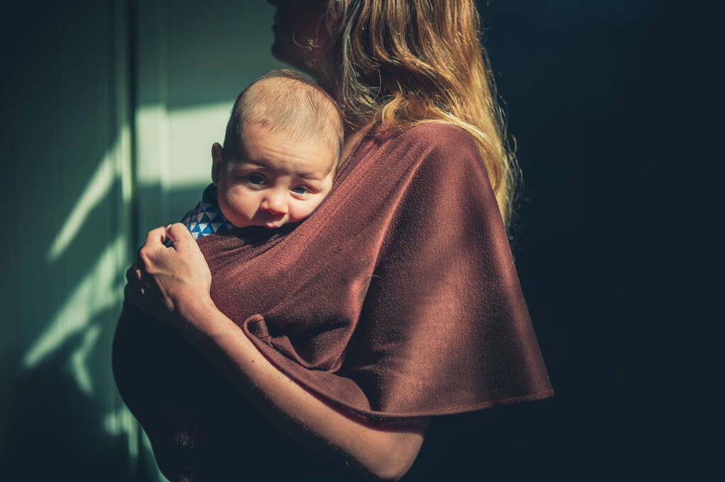 A young mother with her baby in a sling is standing indoors in the sunlight - Photo, Image