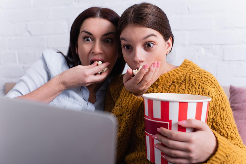 attente tiener meisje en moeder kijken film op laptop en het eten van popcorn in de slaapkamer - Foto, afbeelding