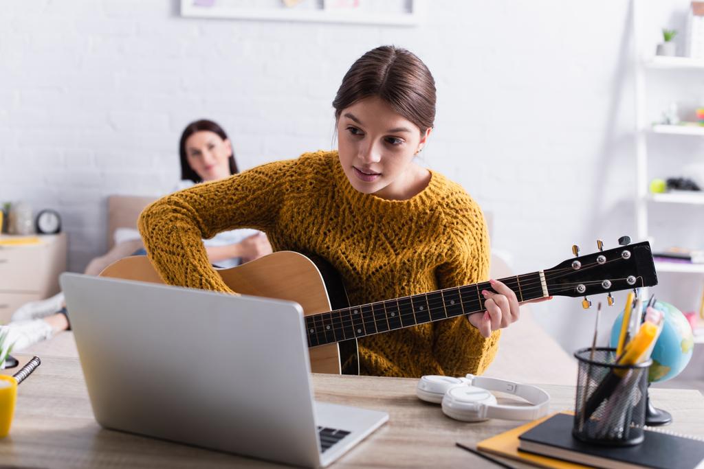 adolescente aprendendo a tocar guitarra acústica perto de laptop e mãe no fundo embaçado  - Foto, Imagem