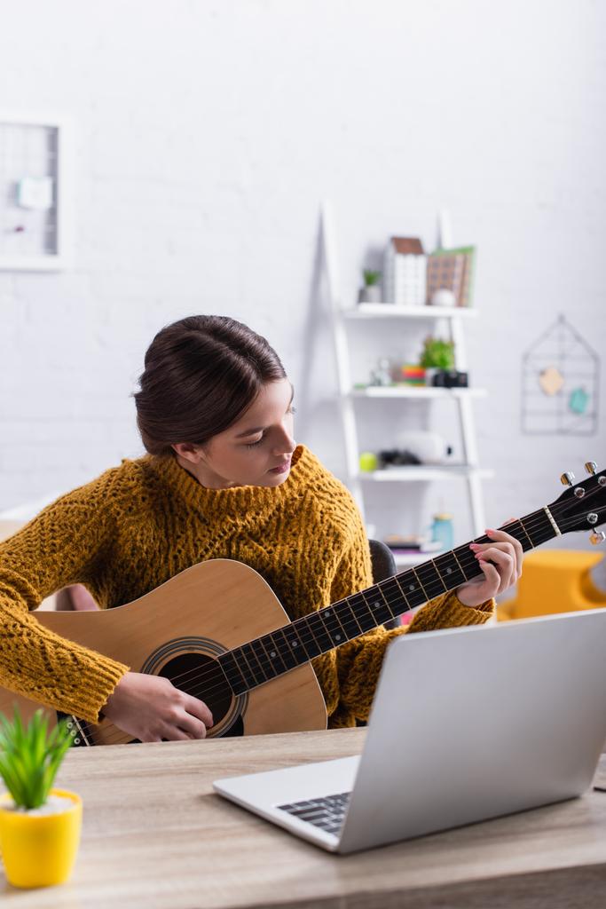 tizenéves lány tanul, hogyan kell játszani akusztikus gitár közelében laptop  - Fotó, kép