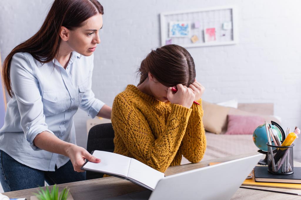 displeased mother holding notebook and looking at crying teenage daughter near laptop - Photo, Image