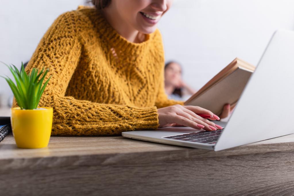 обрезанный вид веселой девочки-подростка, печатающей на клавиатуре ноутбука и держащей книгу - Фото, изображение