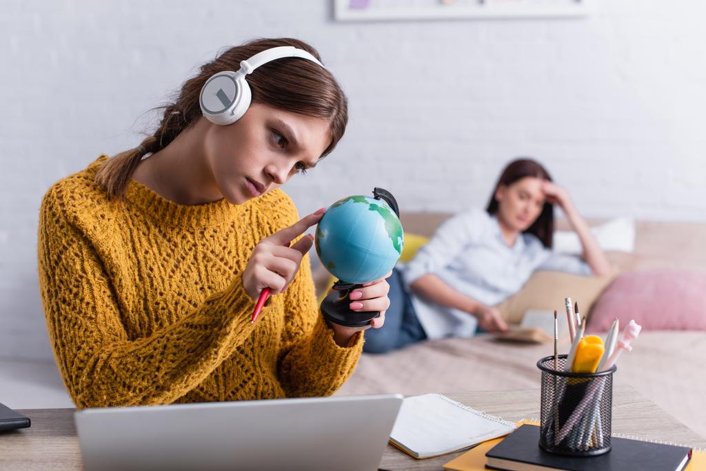triste adolescente in cuffie wireless guardando globo con madre offuscata sullo sfondo  - Foto, immagini