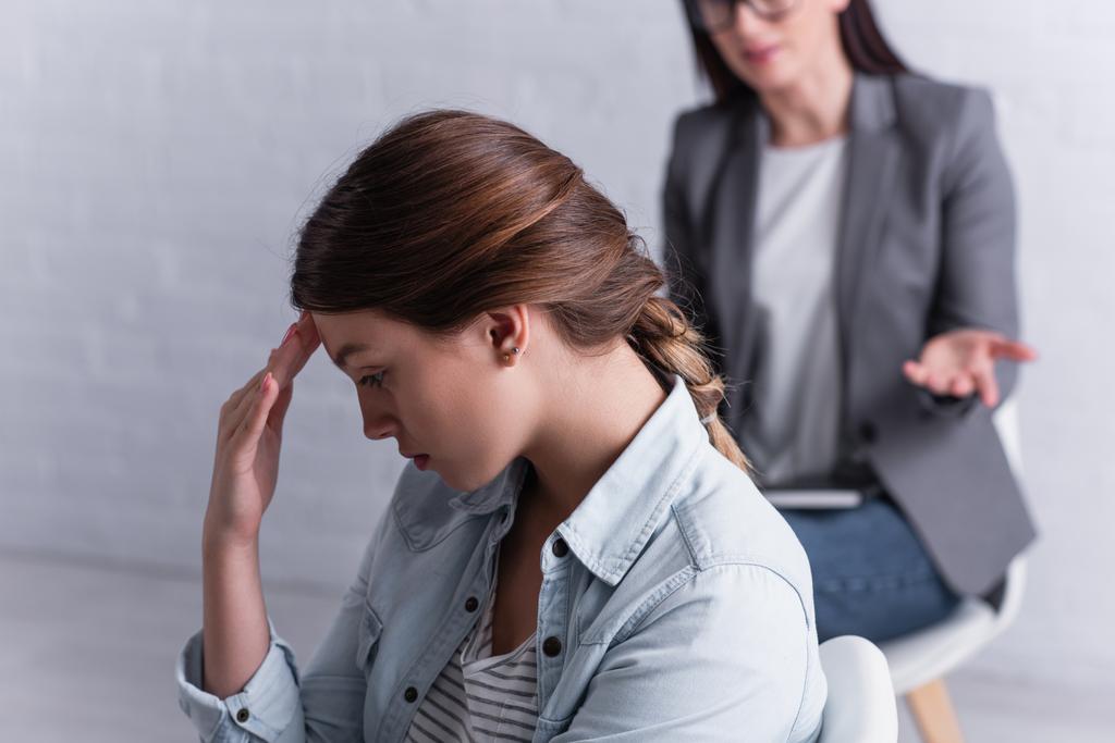 depressed teenage girl looking away near psychologist sitting behind on blurred background  - Photo, Image