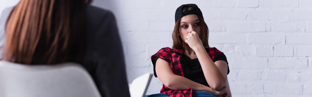 smutný dospívající pacient v čepici sedí a zakrývá ústa poblíž psycholog na rozmazané popředí, prapor - Fotografie, Obrázek