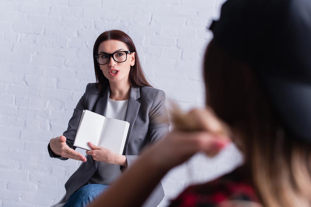 psicólogo en gafas con cuaderno en blanco y gestos mientras mira a un paciente adolescente en primer plano borroso  - Foto, Imagen