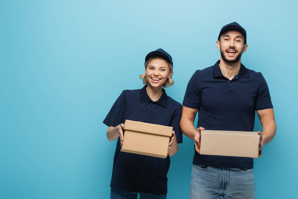 young interracial couriers smiling at camera while holding parcels isolated on blue - Photo, Image