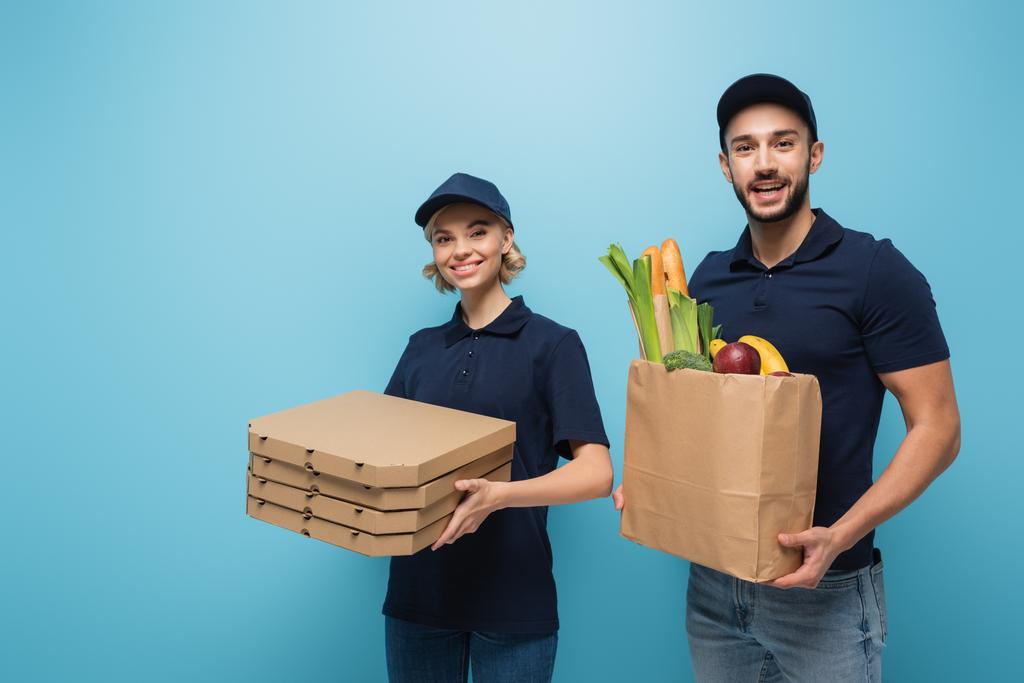 joyful multicultural couriers with pizza boxes and paper bag with food isolated on blue - Photo, Image
