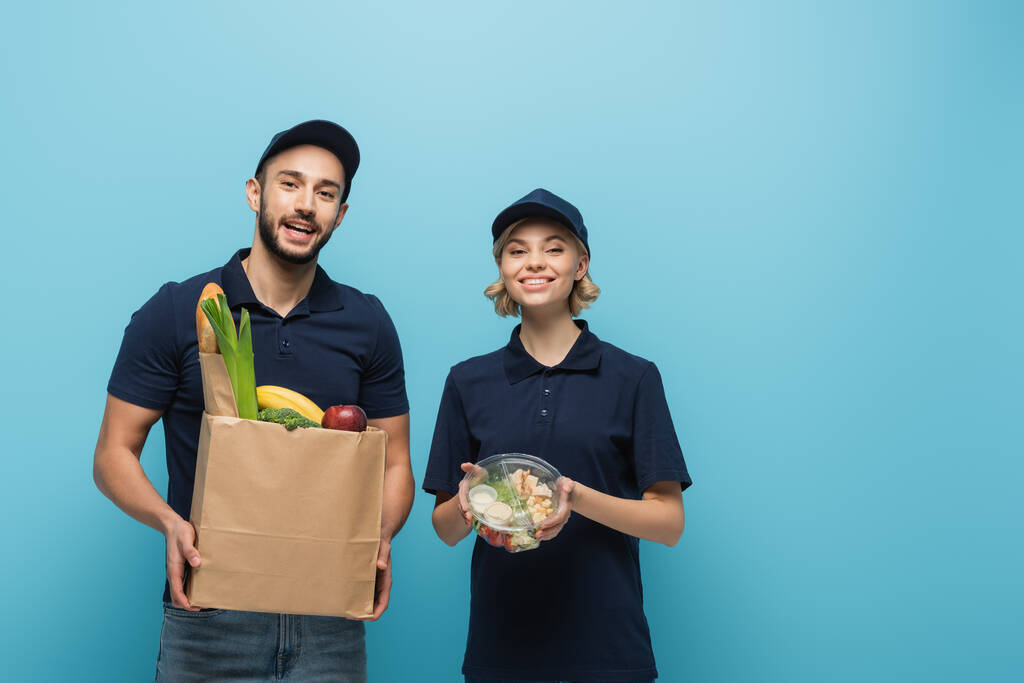 alegres mensajeros interracial sosteniendo paquetes con comida mientras sonríe a la cámara aislada en azul - Foto, imagen