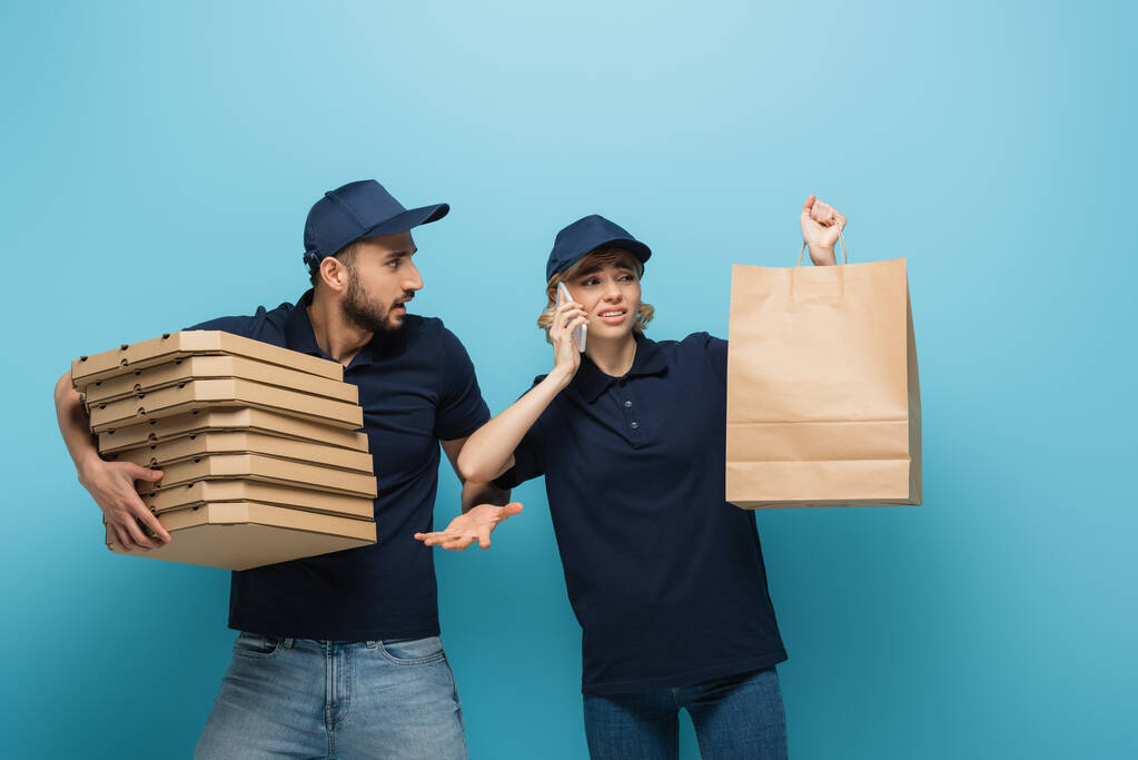 muslim delivery man gesturing while holding pizza boxes near displeased συνάδελφος μιλώντας στο κινητό στο μπλε - Φωτογραφία, εικόνα