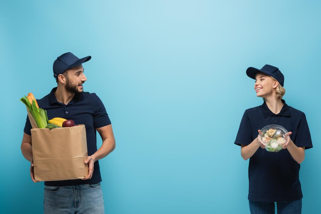 correio multicultural feliz olhando um para o outro enquanto segurando pacotes com alimentos em azul - Foto, Imagem