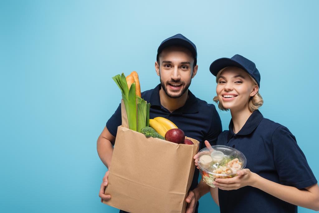 mensageiros multiculturais alegres com alimentos em embalagens sorrindo para a câmera isolada em azul - Foto, Imagem