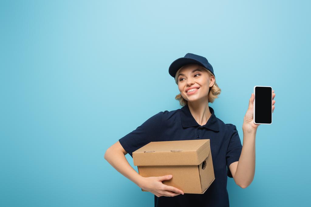 happy courier with carton box and mobile phone with blank screen looking away isolated on blue - Photo, Image