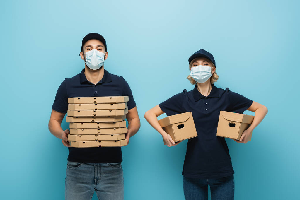 různé kurýři v ochranných maskách drží krabice od pizzy a kartonové balíčky na modré - Fotografie, Obrázek