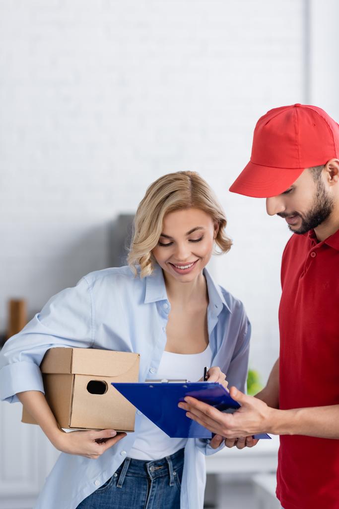 smiling woman signing order on clipboard near arabian delivery man - Photo, Image