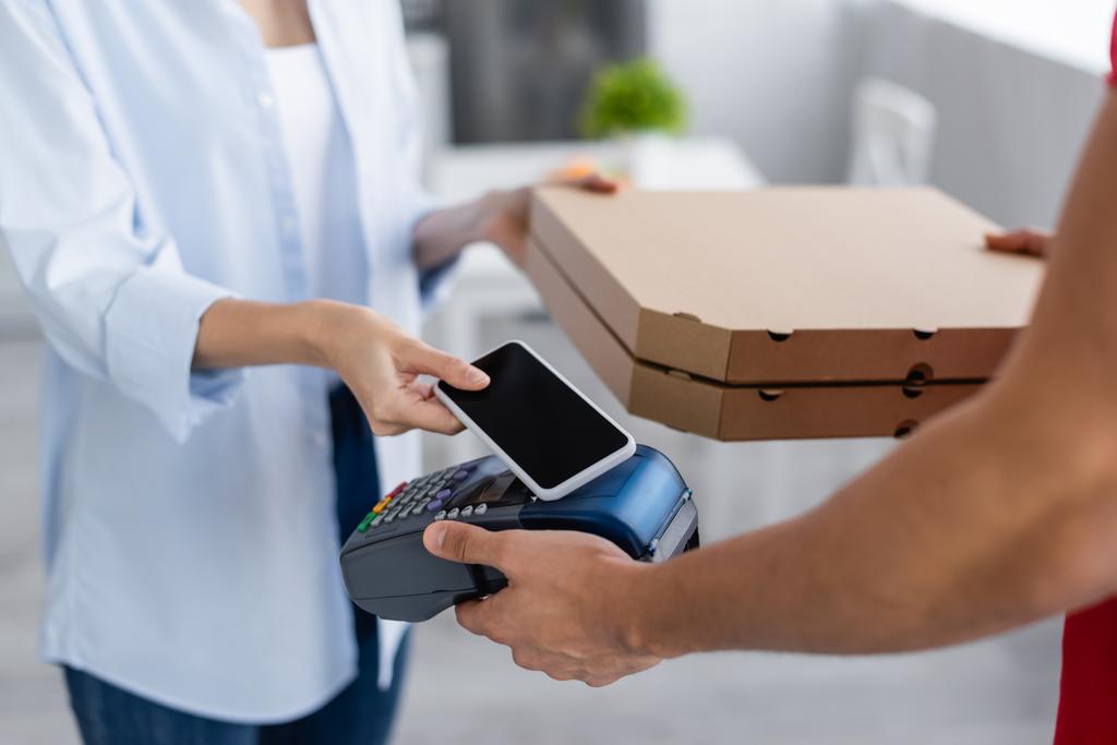 bijgesneden weergave van de levering man met betaalterminal en pizza dozen in de buurt van een vrouw met mobiele telefoon - Foto, afbeelding