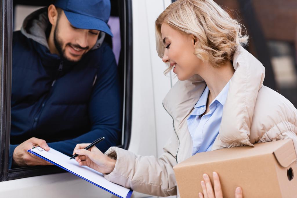 joyful woman signing order while holding parcel near arabian postman in truck on blurred background - Photo, Image