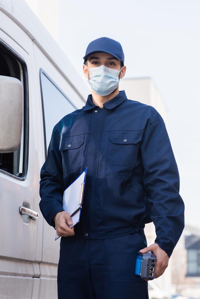 cartero árabe en máscara protectora de pie cerca del coche con terminal de pago y portapapeles - Foto, imagen