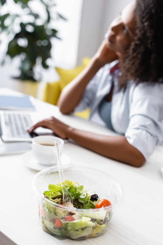 Verse salade in de buurt van Afrikaanse Amerikaanse telewerker met behulp van laptop op wazig achtergrond  - Foto, afbeelding