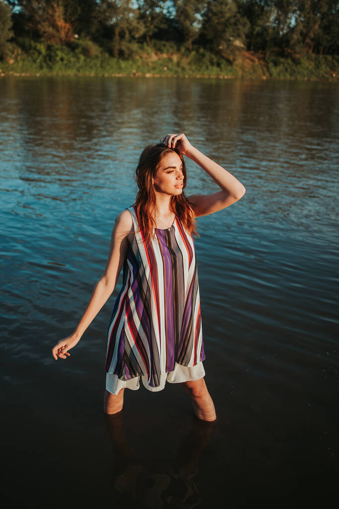 A vertical shot of a young caucasian woman in a striped dress standing in a lake - Фото, изображение