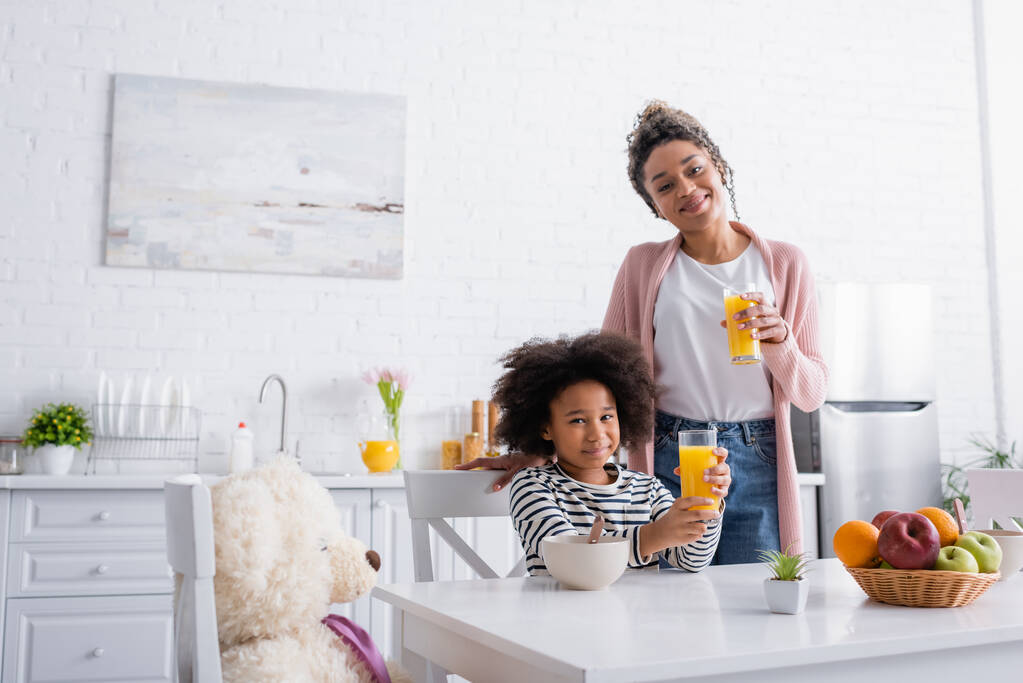 felice afroamericana madre e figlia guardando la fotocamera mentre tiene il succo d'arancia in cucina - Foto, immagini