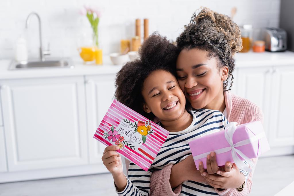 mujer afroamericana emocionada abrazando a hija feliz sosteniendo caja de regalo y tarjeta de día de madres felices - Foto, Imagen