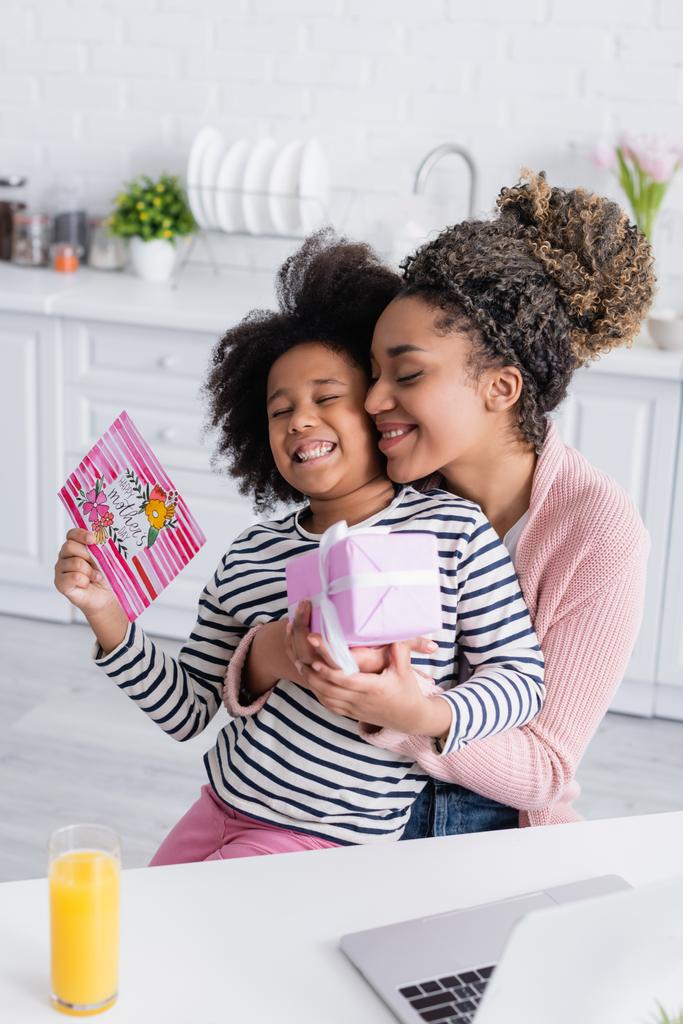 erfreut afrikanisch-amerikanische Frau umarmt Tochter mit Geschenk und glücklicher Muttertagskarte - Foto, Bild