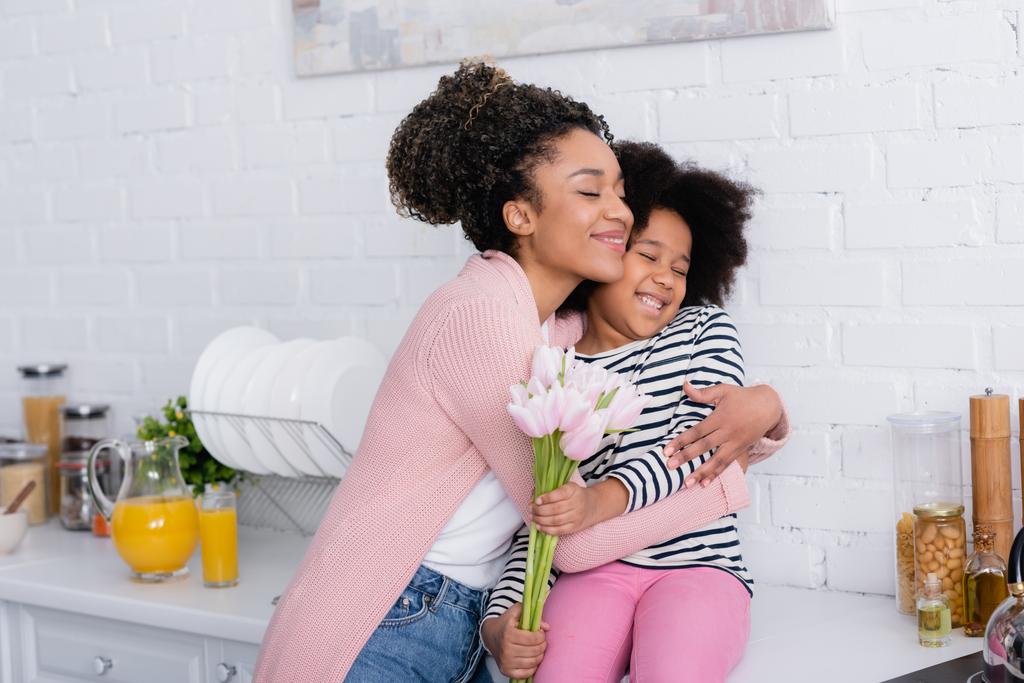 complacida mujer afroamericana abrazando a su hija sentada en el mostrador de la cocina con tulipanes - Foto, imagen