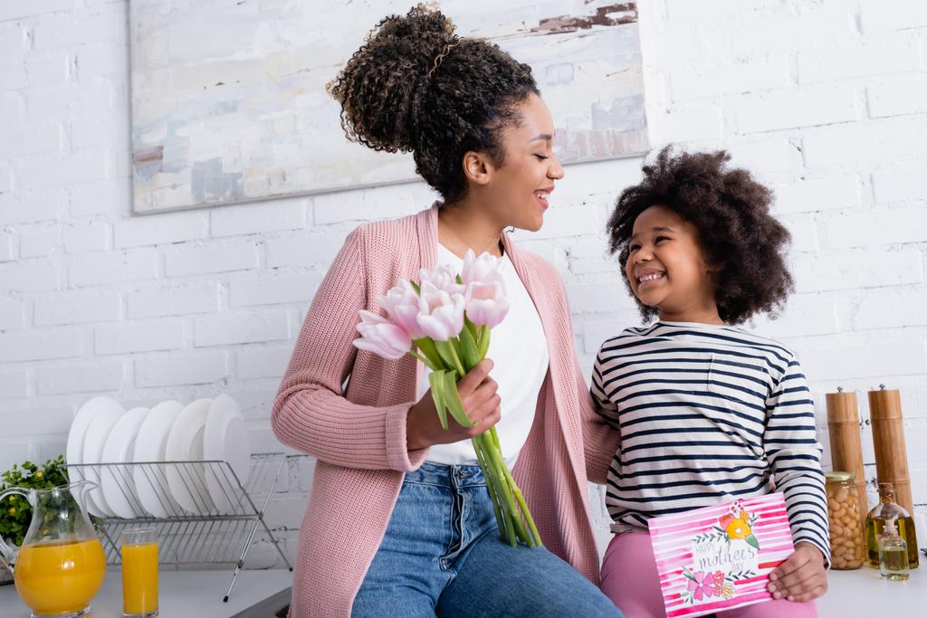 happy african american woman holding tulips near cheerful daughter with happy mothers day card - Photo, Image