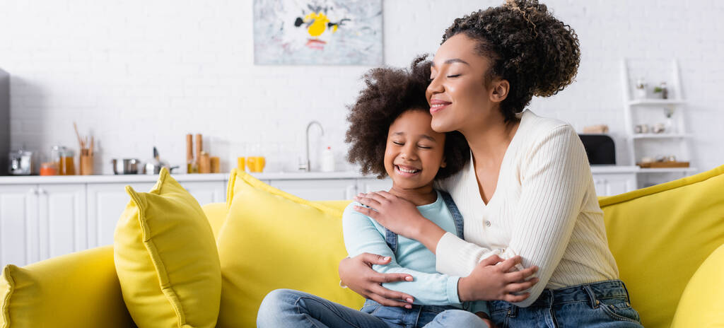 heureux enfant afro-américain et maman embrassant les yeux fermés sur le canapé à la maison, bannière - Photo, image