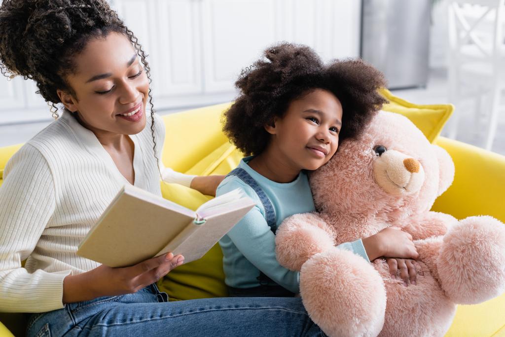 smiling african american woman reading book to happy daughter holding teddy bear - Photo, Image